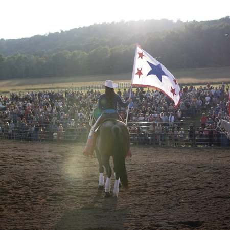 Woman holding a flag pole with right hand while riding a horse. Photo by Kaylin Kavanagh