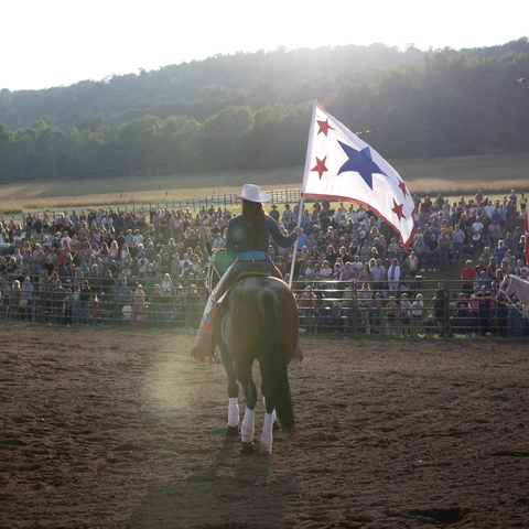 Woman holding a flag pole with right hand while riding a horse. Photo by Kaylin Kavanagh